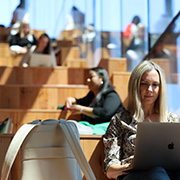 People working on laptops in a modern open workspace with wooden tiered seating, including a blonde woman focused on her laptop in the foreground.