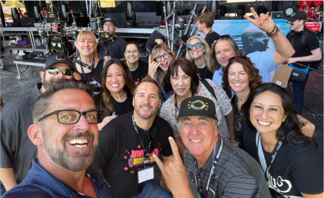 A cheerful group selfie with people smiling and posing on a concert stage setup. Equipment and crew are visible in the background, and everyone appears energetic and excited.