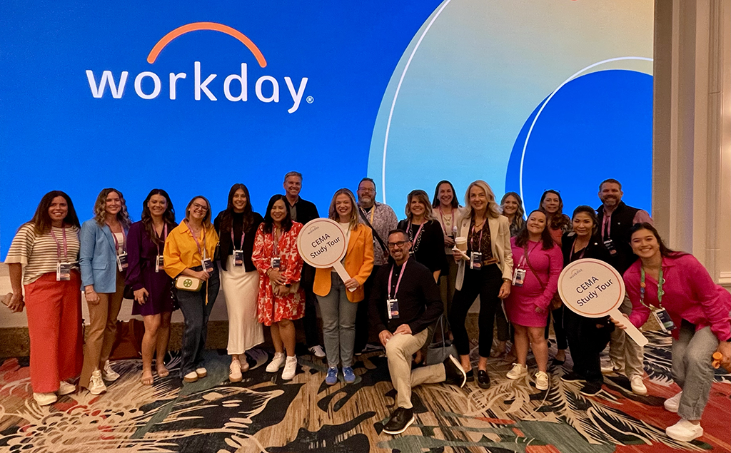 A group of people posing together in front of a large screen displaying the "Workday" logo on a blue background. Two participants are holding "CEMA Study Tour" signs. The group is dressed in vibrant and professional attire, standing on a patterned carpet in a conference setting.